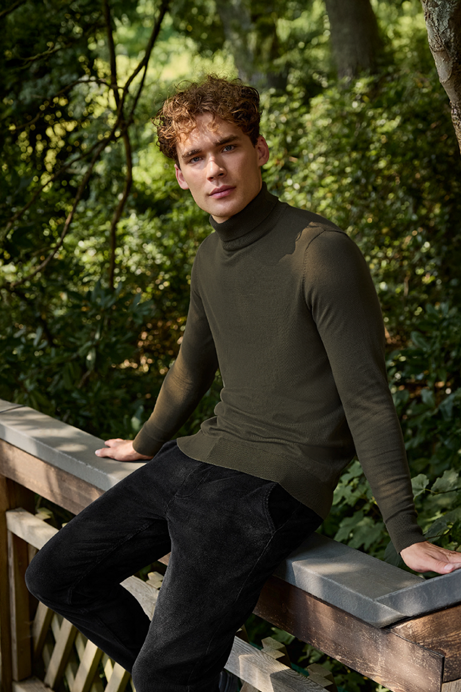 Άνδρας με χακί ζιβάγκο και μαύρο κοτλέ παντελόνι κάθεται σε ξύλινο κάγκελο.