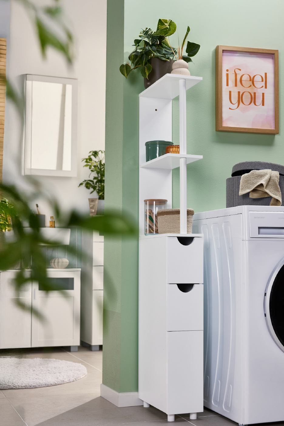 Λευκή ραφιέρα μπάνιου με συρτάρια δίπλα σε πλυντήριο ρούχων, με πράσινο τοίχο και φυτά.