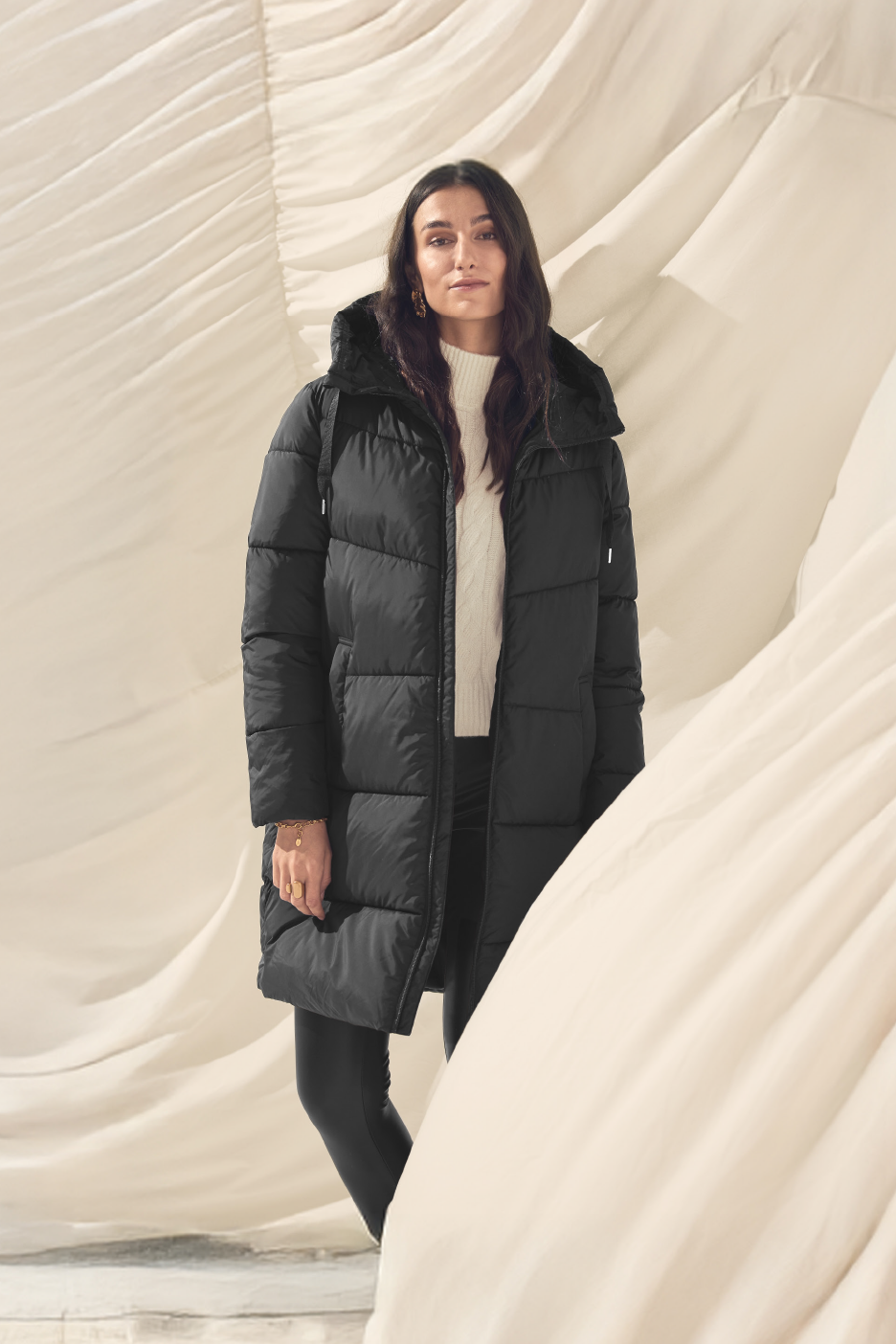 Μια γυναίκα φοράει ένα μακρύ μαύρο φουσκωτό μπουφάν και ένα λευκό πλεκτό πουλόβερ.