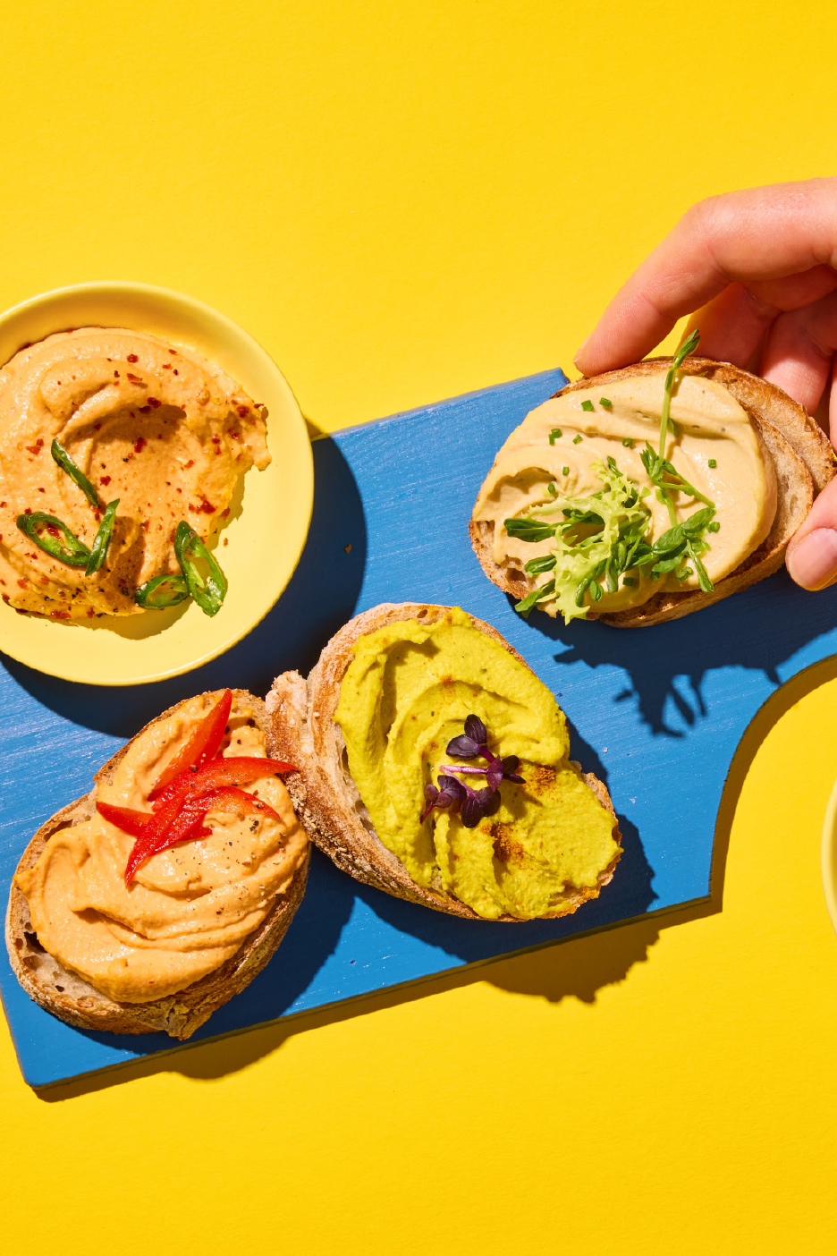 Χούμους σε φέτες ψωμιού και σε μπολ, με διάφορες γαρνιτούρες, σε μπλε και κίτρινο φόντο.