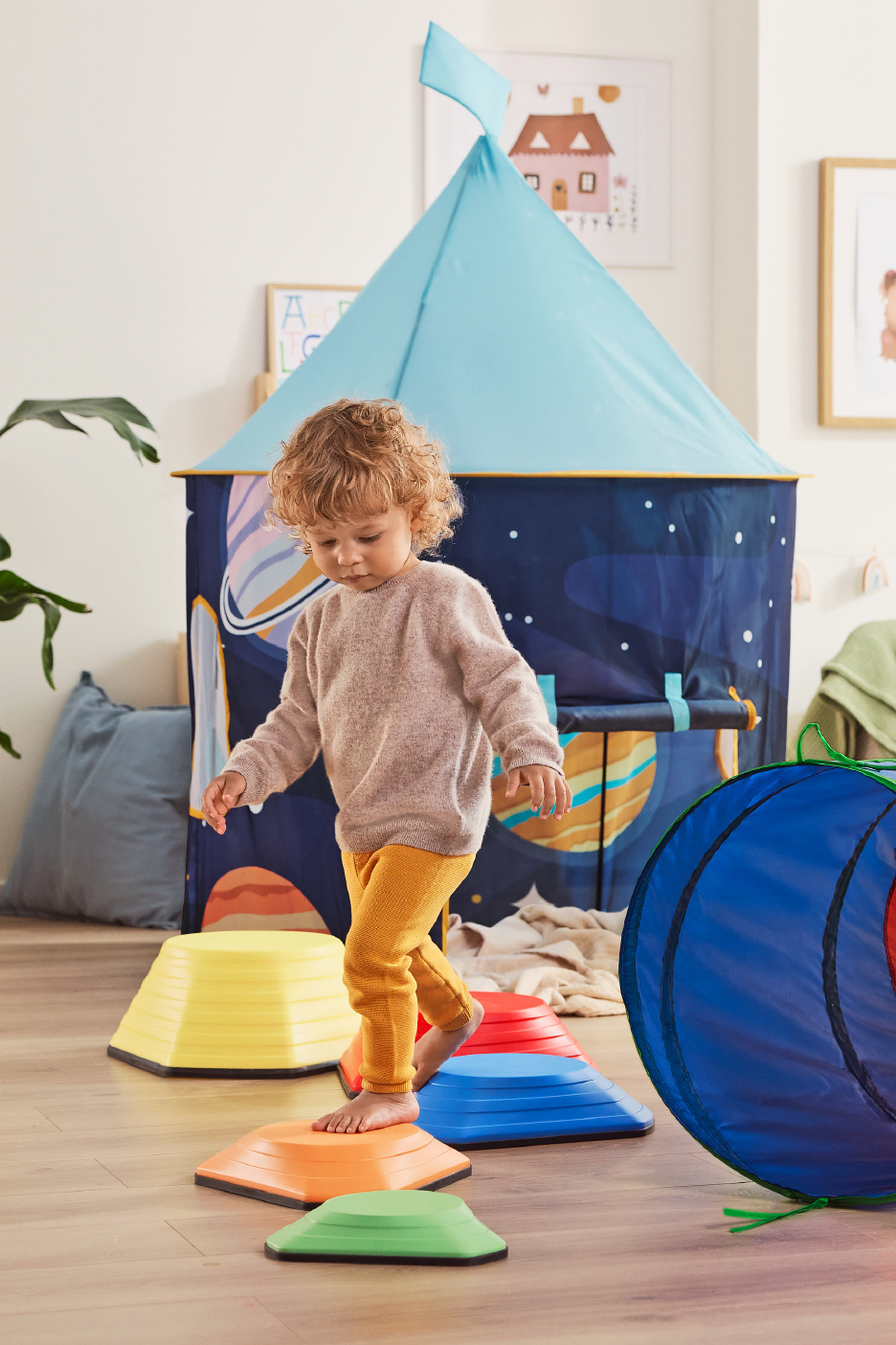 Νήπιο παίζει σε πολύχρωμες πέτρες ισορροπίας μπροστά από μια παιδική σκηνή με θέμα το διάστημα.