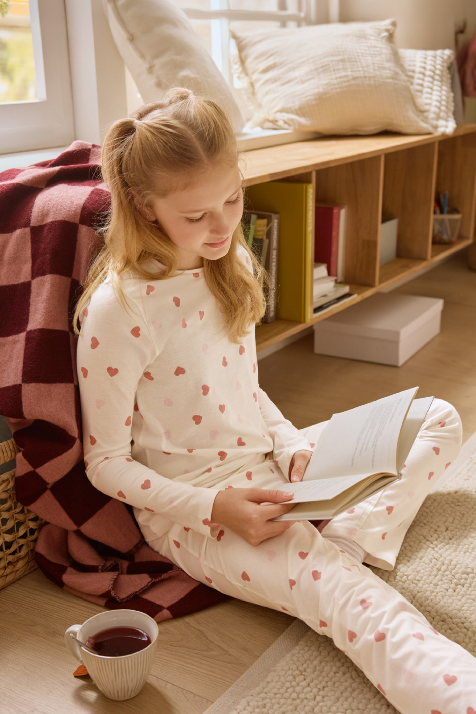 Κορίτσι με πιτζάμες με καρδιές διαβάζει βιβλίο δίπλα σε κουβέρτα και κούπα τσαγιού.