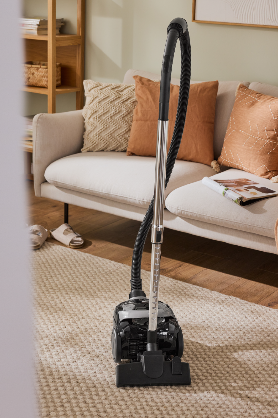 Μαύρη ηλεκτρική σκούπα σε χαλί, με καναπέ και ράφια στο φόντο.