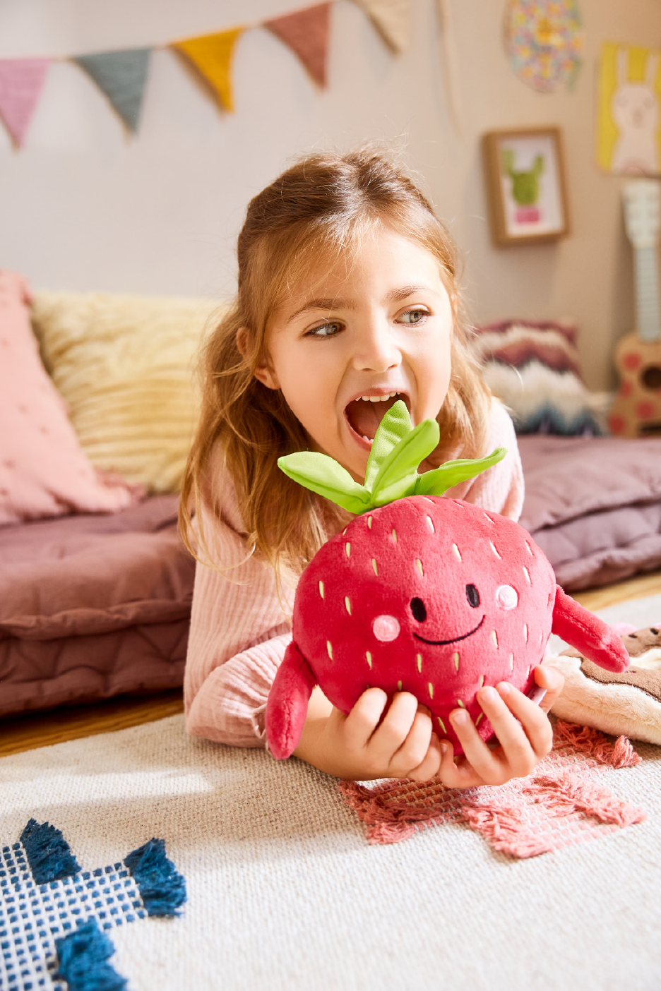 Κορίτσι με λούτρινη φράουλα σε παιδικό δωμάτιο με μαξιλάρια και διακοσμητικά.