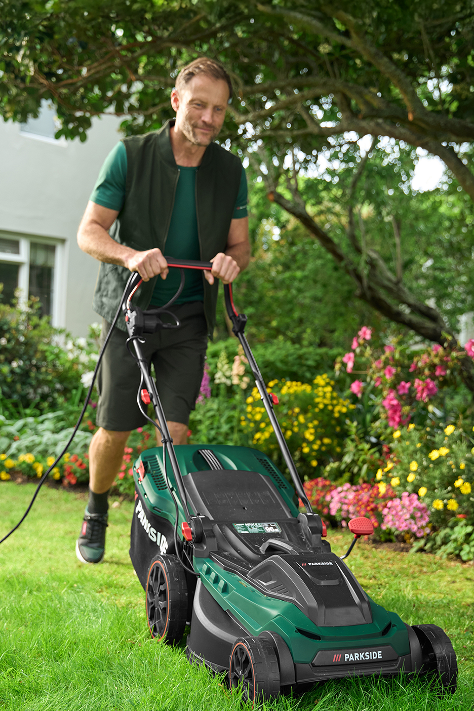 Ένας άντρας σπρώχνει ένα χλοοκοπτικό σε έναν καταπράσινο κήπο με λουλούδια.