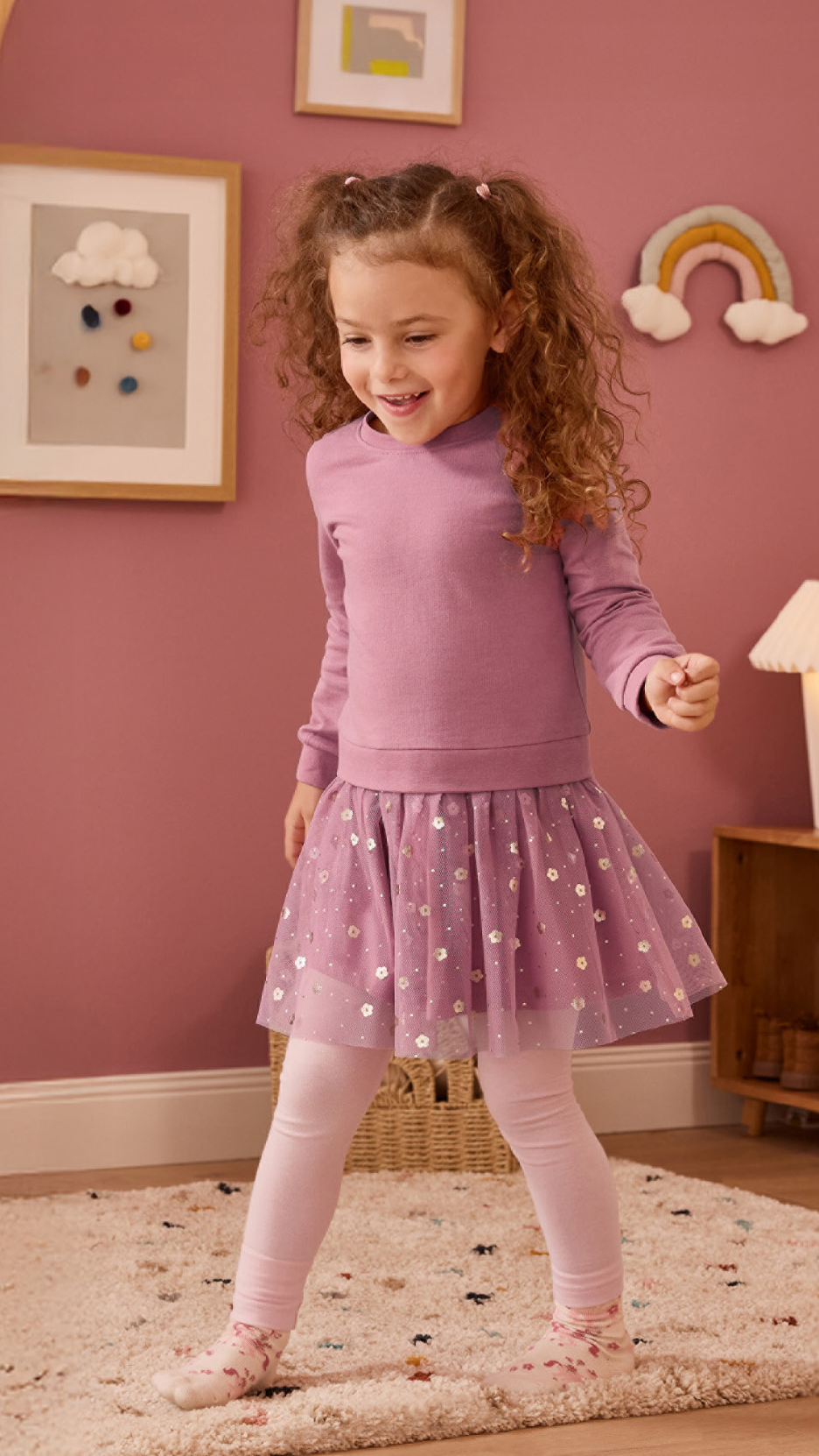 Κορίτσι με ροζ μπλούζα, φούστα με πούλιες και κολάν, χαμογελάει σε παιδικό δωμάτιο.