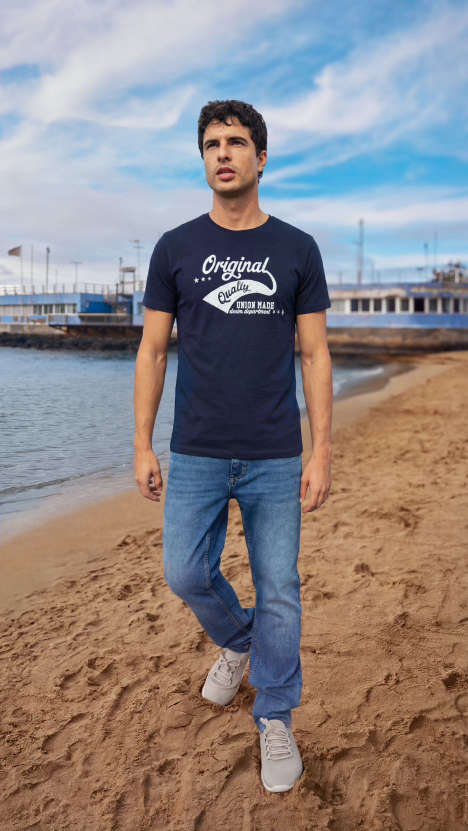 Άνδρας με μπλε τζιν και σκούρο μπλε μπλουζάκι με στάμπα «Original Quality» περπατά στην παραλία.