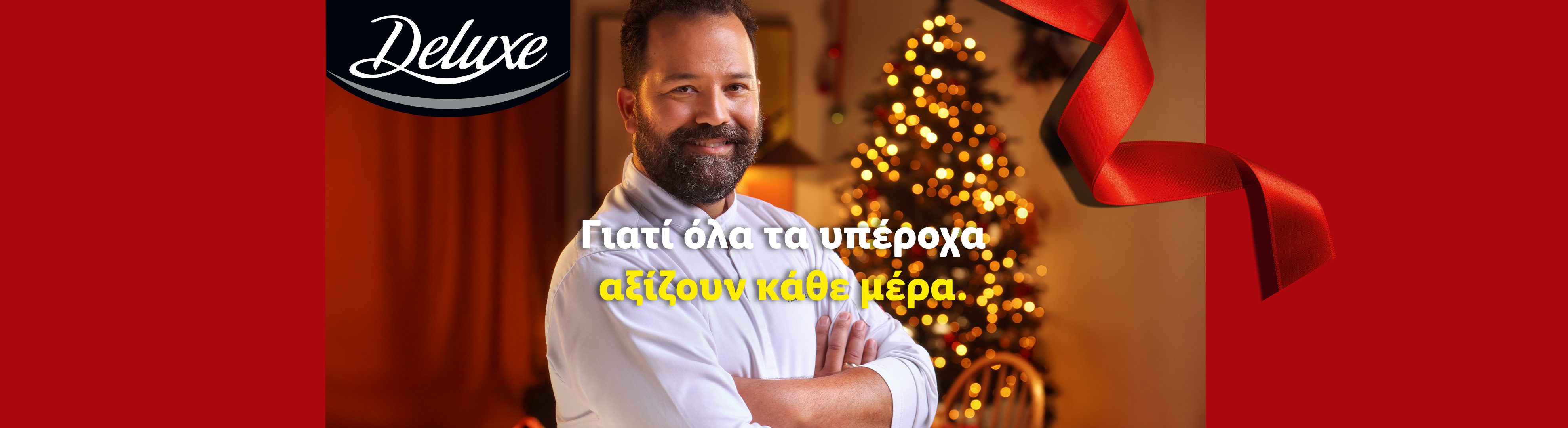 Άνδρας με γενειάδα, φορώντας λευκό πουκάμισο, με φόντο χριστουγεννιάτικο δέντρο και κόκκινη κορδέλα. Λογότυπο Deluxe.